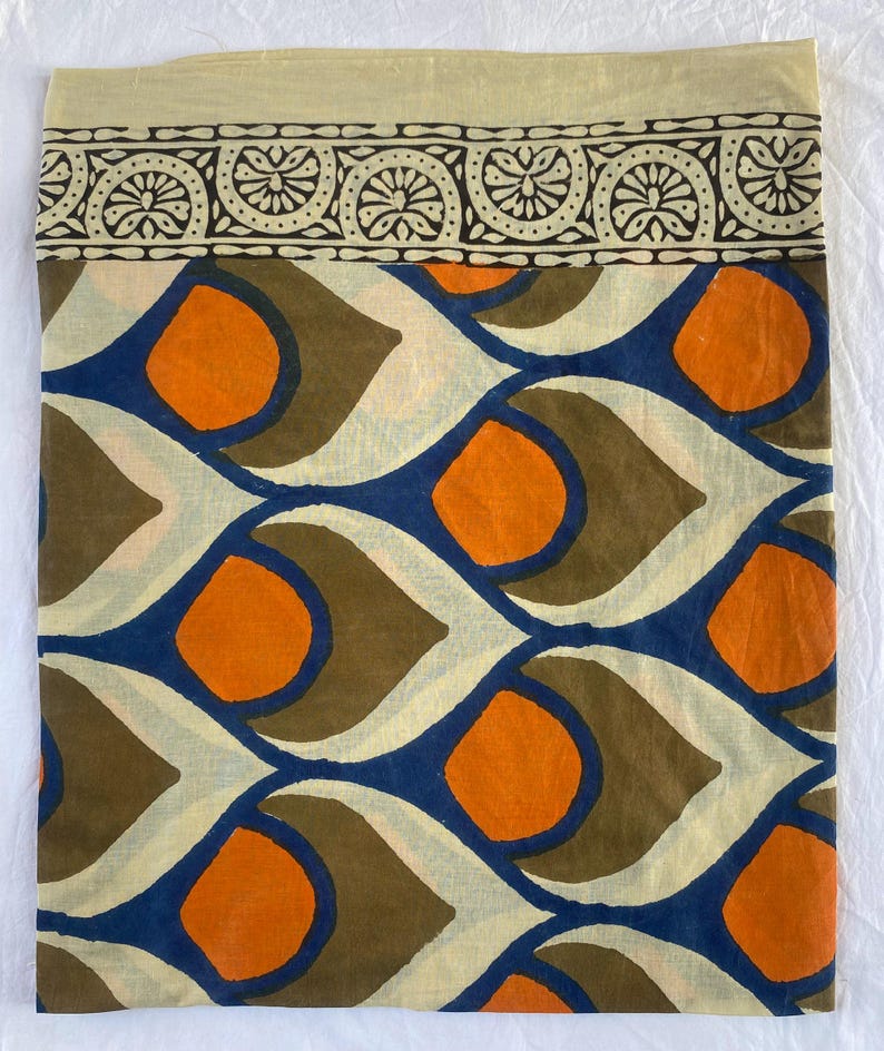 May include: A folded textile with a cream-colored top border featuring a black and cream floral pattern. The main body displays a repeating pattern of orange circles, blue teardrop shapes, and brown and cream elements.