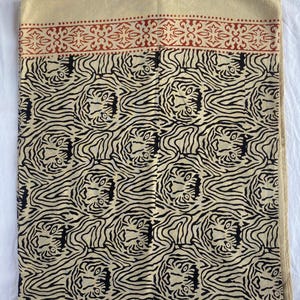 May include: A beige textile with a repeating black tiger stripe pattern. The top border features a red floral design. The fabric is folded, showing the pattern and color variations. The textile is likely a scarf or shawl.