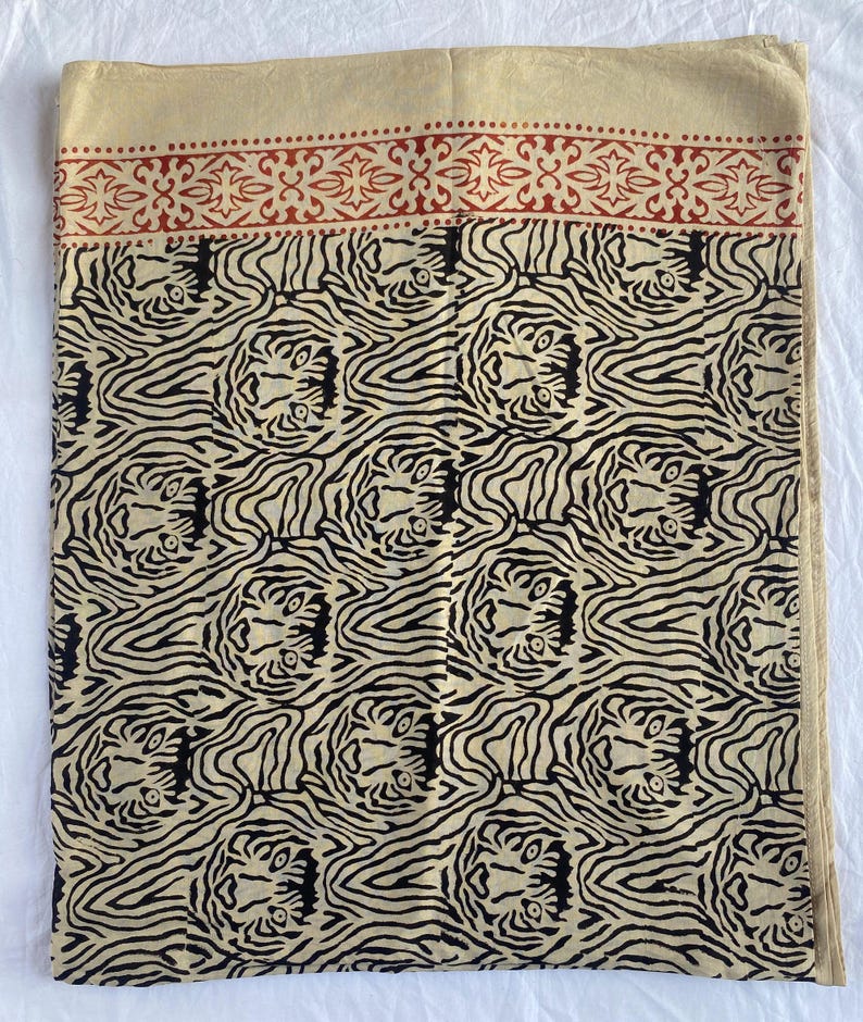 May include: A beige textile with a repeating black tiger stripe pattern. The top border features a red floral design. The fabric is folded, showing the pattern and color variations. The textile is likely a scarf or shawl.