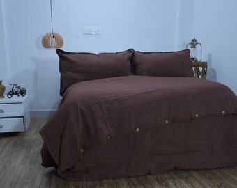 May include: A brown linen duvet cover with button fastenings. The duvet cover is on a bed with two matching pillowcases. The bed is in a bedroom with a white bedside table and a wooden lamp hanging from the ceiling.