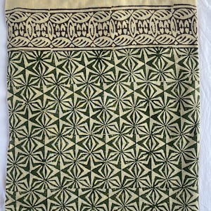 May include: A folded textile with a cream-colored top border and a dark brown patterned design. Below, a repeating geometric pattern in dark green and cream covers the majority of the fabric. The design features star-like shapes.