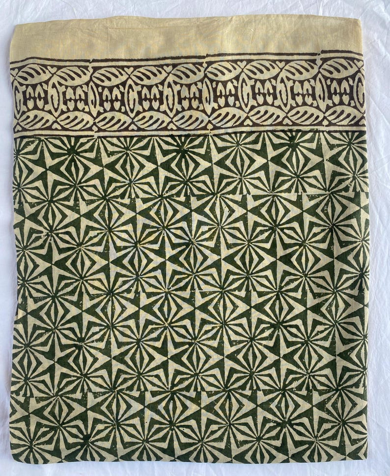 May include: A folded textile with a cream-colored top border and a dark brown patterned design. Below, a repeating geometric pattern in dark green and cream covers the majority of the fabric. The design features star-like shapes.
