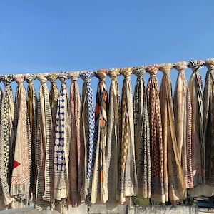 May include: A collection of colorful, patterned scarves hanging on a metal rod against a clear blue sky. The scarves feature various designs, including geometric shapes, stripes, and floral motifs, in shades of red, blue, and beige.