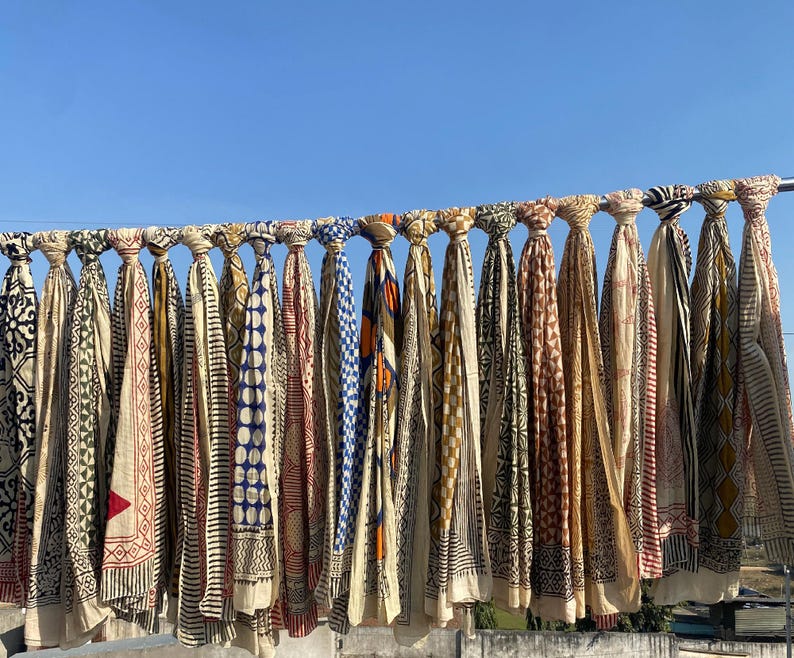 May include: A collection of colorful, patterned scarves hanging on a metal rod against a clear blue sky. The scarves feature various designs, including geometric shapes, stripes, and floral motifs, in shades of red, blue, and beige.