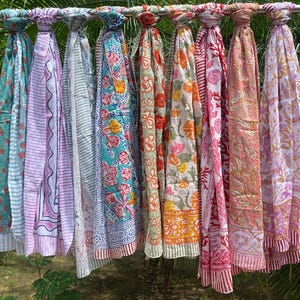 Peut inclure: Une collection d'écharpes colorées à motifs, suspendues à une barre métallique. Les écharpes présentent divers motifs floraux et géométriques dans des tons de bleu, rose, orange et vert. Le tissu semble léger, idéal pour l'été.
