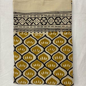 Può includere: Tessuto piegato con parte superiore color crema e motivo. Il motivo presenta forme geometriche nere e motivi a forma di goccia giallo senape su sfondo crema. Il tessuto è piegato ordinatamente, mostrando il design.