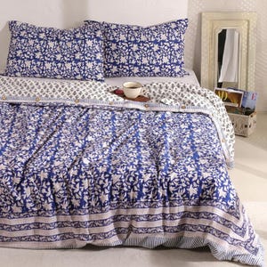Op de afbeelding: Een blauw en wit bloemmotief beddengoed set. De set bevat twee kussenslopen, een dekbedovertrek en een decoratieve rand. Een kopje thee staat op het bed. Het beddengoed heeft een bohemien stijl.