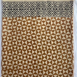 May include: A folded textile featuring a geometric pattern in brown and cream. The top section has a black and cream geometric border. The lower section has a repeating pattern of brown triangles on a cream background. The textile is likely cotton or linen.