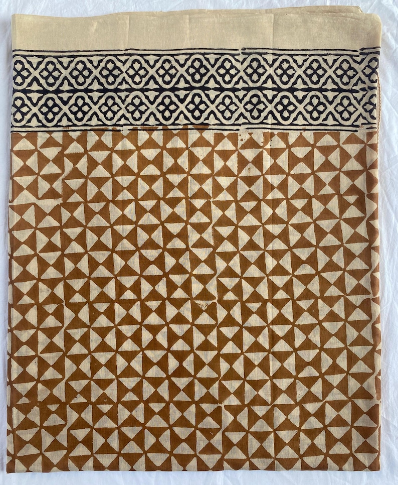 May include: A folded textile featuring a geometric pattern in brown and cream. The top section has a black and cream geometric border. The lower section has a repeating pattern of brown triangles on a cream background. The textile is likely cotton or linen.