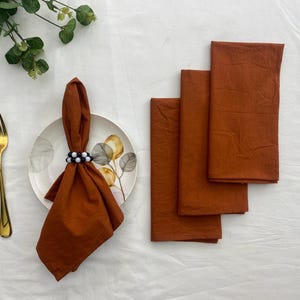 Puede incluir: Un conjunto de servilletas de lino color óxido, dobladas y apiladas, junto con un servilletero a juego con cuentas blancas y negras. También hay un tenedor dorado y un plato con un diseño floral, lo que sugiere un entorno de comedor.