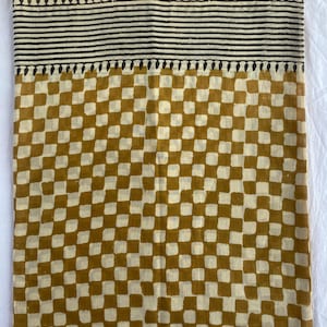 May include: A textile with a cream-colored top border featuring a black leaf-like pattern. Below are horizontal black stripes, followed by a mustard yellow and cream checkered pattern. The fabric is likely cotton or linen.