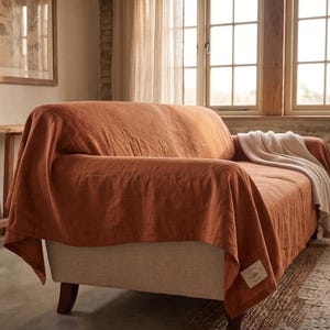 May include: A rust-colored linen sofa cover draped over a light beige sofa, featuring a small rectangular white fabric label with text on the lower corner and a folded white throw blanket resting on the seat cushion of the sofa.