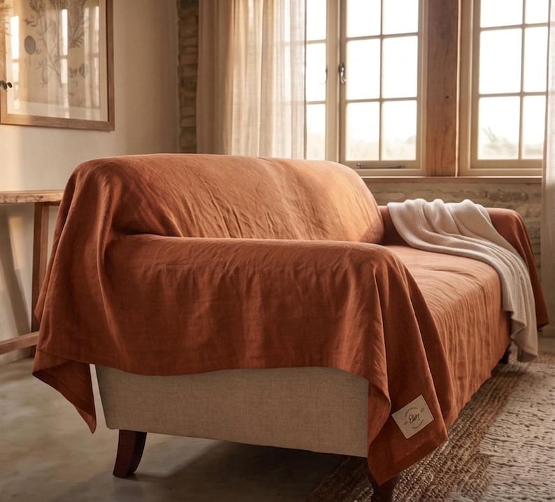 May include: A rust-colored linen sofa cover draped over a light beige sofa, featuring a small rectangular white fabric label with text on the lower corner and a folded white throw blanket resting on the seat cushion of the sofa.