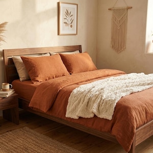 May include: A wooden bed frame with rust-colored bedding and a cream-colored throw blanket. The room features a wooden nightstand, a framed botanical print, and a macrame wall hanging. A vase of dried pampas grass sits on the nightstand.