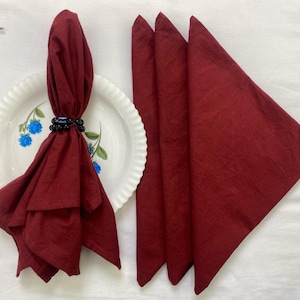 Könnte beinhalten: Ein Set aus weinroten Leinen-Servietten, eine gefaltet und von einem schwarzen Perlen-Serviettenring gehalten, und drei dreieckig gefaltet. Die Servietten liegen auf einem weißen Teller mit floralem Design.