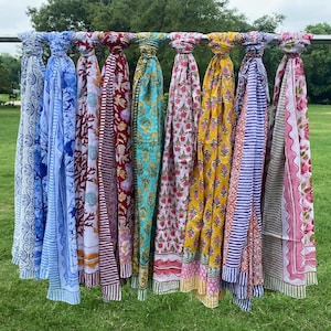 May include: A collection of nine rectangular cotton scarves hanging from a horizontal metal bar. Each scarf features a distinct block-printed pattern in various colors including blue, red, yellow, turquoise, pink, and white with floral and geometric motifs.