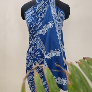 Peut inclure: Un sarong en coton bleu avec un imprimé de poissons et de cachemire blancs. Le sarong est drapé sur un mannequin et est noué autour du cou.
