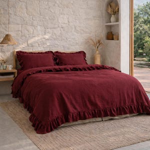 May include: A bedroom with a bed dressed in deep red bedding, including a duvet cover and pillowcases, all with a ruffled trim. The bed rests on a woven jute rug. The room has a neutral colour scheme, with a stone wall and natural light.