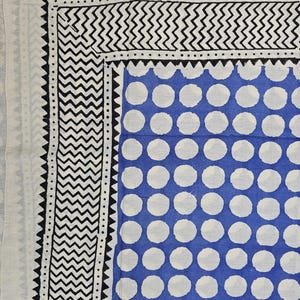 Op de afbeelding: Een witte stof met een blauw en wit stippenpatroon. De stof heeft een zwarte en witte rand met een zigzagpatroon.