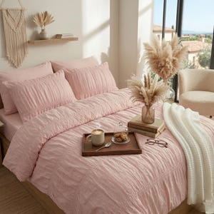 Peut inclure: Un lit avec une couette rose clair et des taies d'oreiller assorties. Un plateau en bois contient une tasse de café, des biscuits et une cuillère. Une couverture blanche est drapée sur le lit. Des plantes séchées décoratives et une tenture murale en macramé sont également visibles.