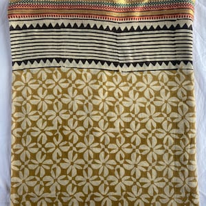 May include: A folded textile featuring a floral pattern in shades of beige and gold. The top section has horizontal black stripes and a decorative border with red, green, and gold accents. The fabric is likely cotton or a similar material.