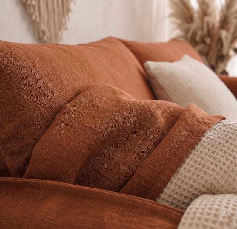 May include: A rust-colored sofa with a textured fabric finish, featuring a matching throw blanket draped over the armrest and a cream-colored waffle-knit pillow positioned against the back cushions, showcasing a soft and woven material appearance.