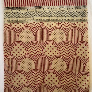 May include: A beige textile with a repeating pattern of red arches, zigzags, and dots. The top border features vertical red stripes and a black and white geometric design. The fabric has a handmade, block-printed appearance.