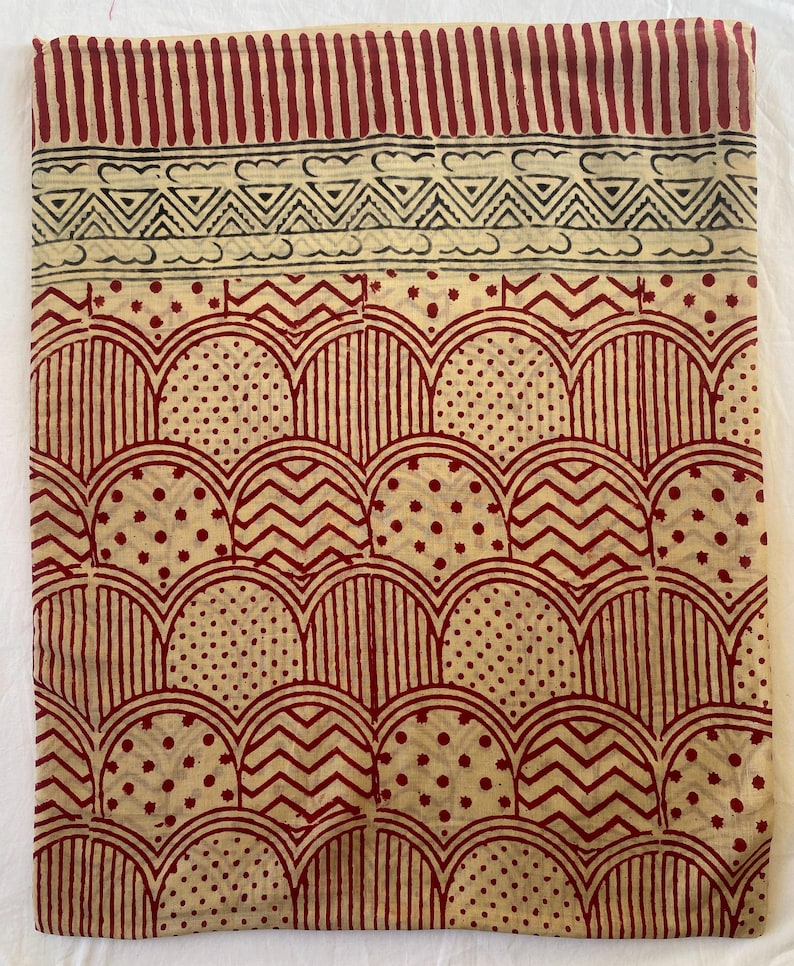 May include: A beige textile with a repeating pattern of red arches, zigzags, and dots. The top border features vertical red stripes and a black and white geometric design. The fabric has a handmade, block-printed appearance.