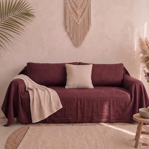 May include: A deep burgundy sofa covered with a textured fabric, featuring a single cream-coloured square throw pillow centred on the back cushions and a light beige waffle-knit throw blanket draped over the left arm and seat of the sofa.
