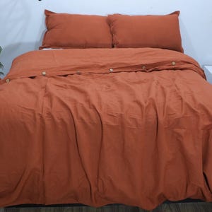 Puede incluir: Una funda nórdica de lino de color óxido con cierre de botones. La funda nórdica está en una cama con dos fundas de almohada a juego.