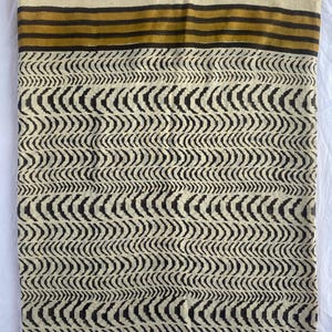 May include: A folded textile with a cream-colored background and a repeating black wave pattern. The top edge features horizontal stripes in brown and gold. The fabric appears to be cotton or a similar natural fiber.