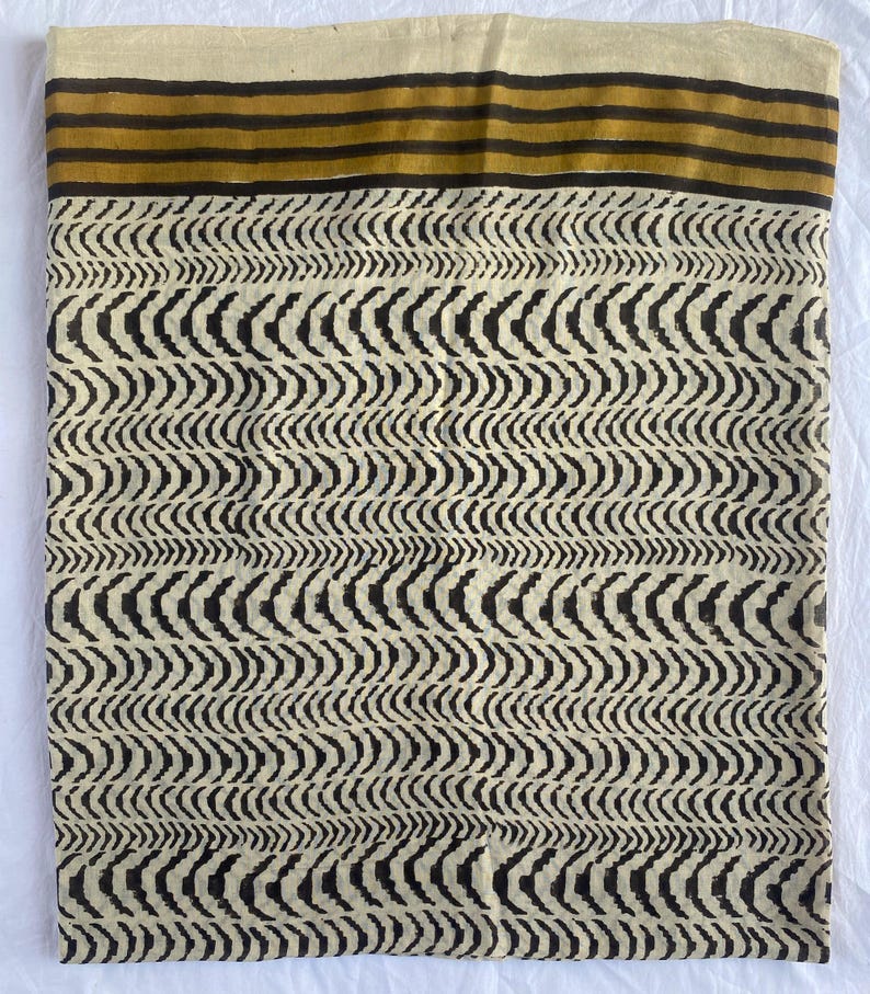 May include: A folded textile with a cream-colored background and a repeating black wave pattern. The top edge features horizontal stripes in brown and gold. The fabric appears to be cotton or a similar natural fiber.