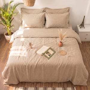 May include: A beige linen bedding set featuring a duvet cover and two matching pillowcases. The bed is dressed in a neutral-toned fabric with a visible weave texture. An open book, a small wooden hand sculpture, and a dried plant are on the bed.