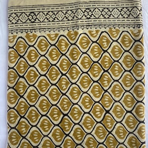 May include: Folded fabric with a repeating pattern of olive green leaf-like shapes outlined in dark brown. The top edge features a decorative border with geometric designs in brown against a beige background. The fabric is likely cotton or linen.