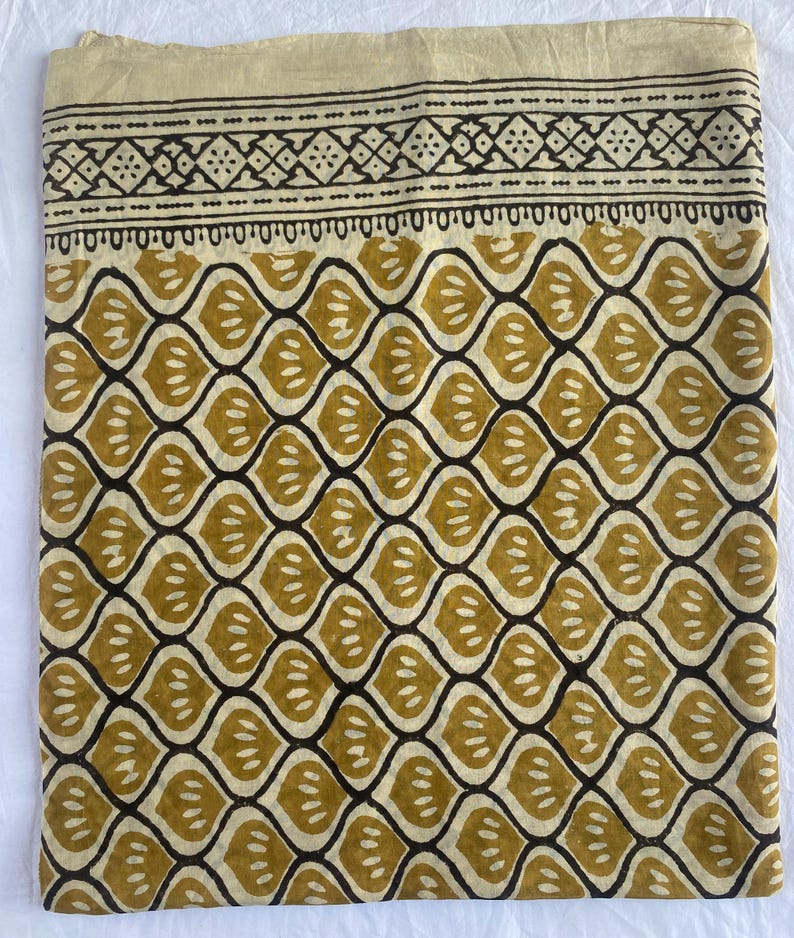 May include: Folded fabric with a repeating pattern of olive green leaf-like shapes outlined in dark brown. The top edge features a decorative border with geometric designs in brown against a beige background. The fabric is likely cotton or linen.