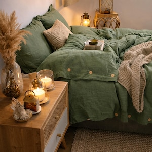 May include: A bedroom scene featuring a bed with olive green bedding, including a duvet cover with wooden button details and matching pillowcases. A beige throw blanket and a striped pillow add texture. A wooden nightstand with a woven drawer front holds candles and decorative items.