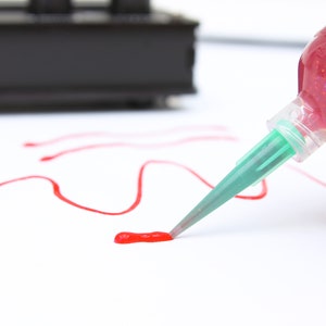 Può includere: Una bottiglia di colla glitterata rossa con un beccuccio verde viene utilizzata per disegnare una linea rossa su una superficie bianca.