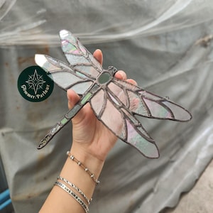 Pode incluir: Uma libélula de vitral com asas iridescentes e corpo verde. A libélula é segurada por uma mão. A libélula tem um design detalhado e é feita de várias peças de vidro. O logotipo "Glimmer Parlour" é visível.