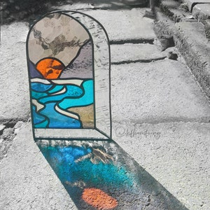 Puede incluir: Una ventana de vitral que representa una puesta de sol sobre una masa de agua. La ventana está enmarcada en negro y tiene una superficie de vidrio texturizado. Los colores de la ventana son naranja, azul y blanco.