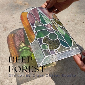 Puede incluir: Un kit de bricolaje de vitrales con un diseño geométrico en colores verde, morado y naranja. El kit se titula "DEEP FOREST" y es de "Glass Glitter Studio".
