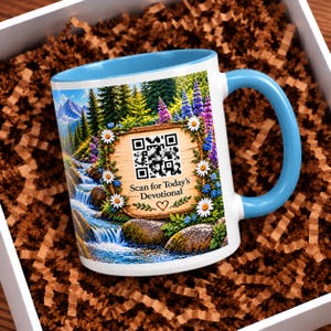 May include: A white ceramic mug with a light blue handle and interior rim. The exterior features a landscape illustration of a mountain stream, wildflowers, and a wooden sign with a QR code and the text Scan for Today's Devotional.