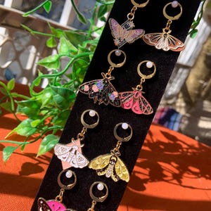 May include: A collection of gold-colored dangle earrings featuring colorful moth designs. Each earring has a small pearl accent and a lever-back closure. The moths are in various colors, including pink, purple, yellow, and black, with intricate details.