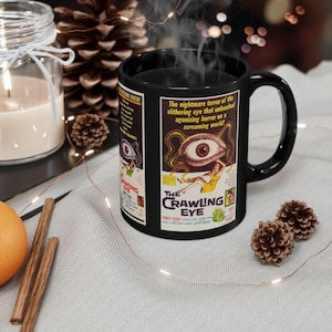 May include: Black ceramic mug with a vintage movie poster design for "The Crawling Eye." The poster art includes an eye, a woman, and the text "The Crawling Eye." The mug has a black handle and is filled with a steaming beverage.