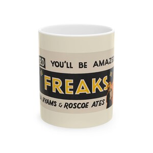 May include: A white ceramic mug featuring a vintage-style graphic design with the text "YOU'LL BE AMAZED" and "FREAKS" in bold yellow and black lettering. The design also includes the names "LEILA HYAMS & ROSCOE ATES" printed in black.