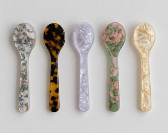May include: Five small spoons arranged in a row, each featuring a distinct marbled or tortoiseshell pattern. The colors include a blue and white mix, a classic brown and black tortoiseshell, a light lavender, a green and pink blend, and a cream.