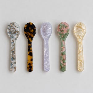 May include: Five small spoons arranged in a row, each featuring a distinct marbled or tortoiseshell pattern. The colors include a blue and white mix, a dark brown tortoiseshell, a light lavender, a green and pink blend, and a translucent cream.
