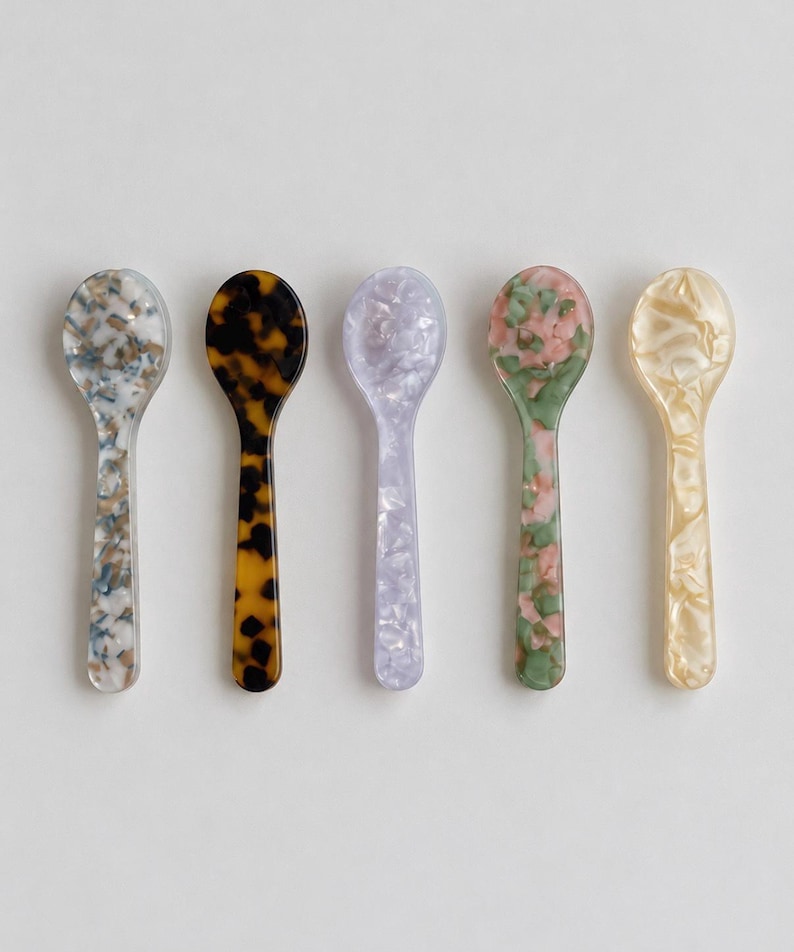 May include: Five small spoons arranged in a row, each featuring a distinct marbled or tortoiseshell pattern. The colors include a blue and white mix, a dark brown tortoiseshell, a light lavender, a green and pink blend, and a translucent cream.