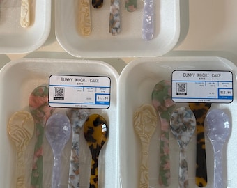Pode incluir: Uma coleção de colheres pequenas com vários padrões de mármore e tartaruga em tons de creme, lavanda, verde e castanho. As colheres estão dispostas em tabuleiros de espuma branca, cada um com uma etiqueta que diz BUNNY MOCHI CAKE.