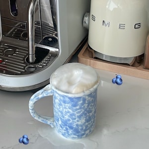 May include: A blue and white speckled mug filled with a creamy latte. The mug is sitting on a white countertop with a few blueberries scattered around it.