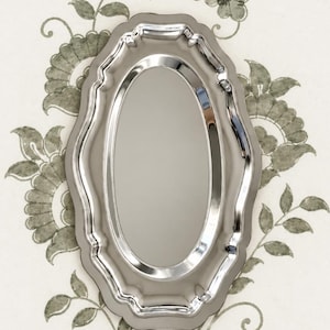 May include: A polished silver-toned serving tray with an oval center and a scalloped, decorative rim. The tray features a reflective, metallic surface with smooth, curved edges and a symmetrical, elongated shape designed for display or service.